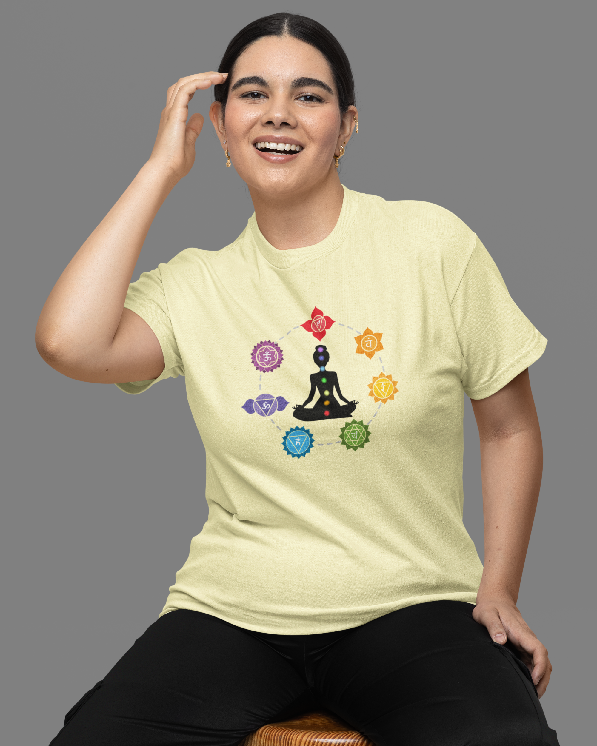 Person wearing a light yellow t-shirt with a colorful design, sitting on a wooden stool against a gray background.