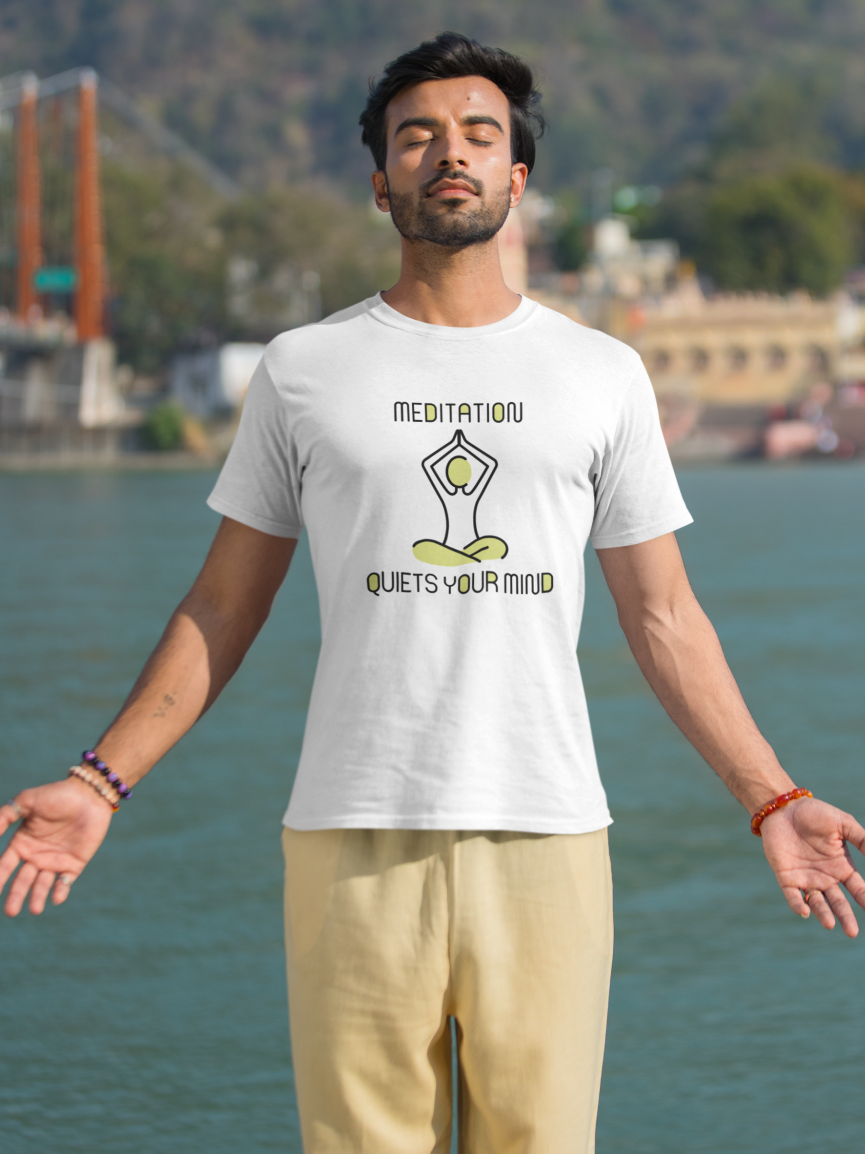Man wearing a t-shirt with 'Meditation quiets your mind' design by a body of water.