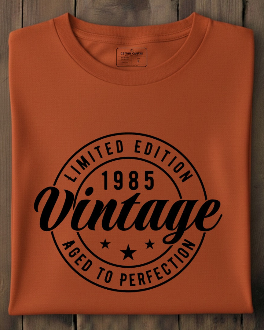 Folded orange t-shirt with black 'Limited Edition 1985 Vintage Aged to Perfection' text on a wooden surface