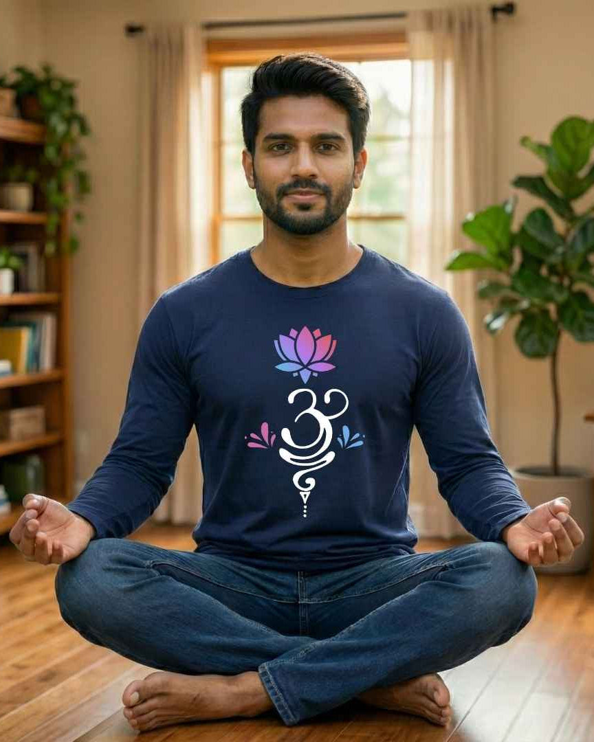 Man sitting in a meditative pose wearing a navy blue shirt with a symbol design in a room with plants and books.